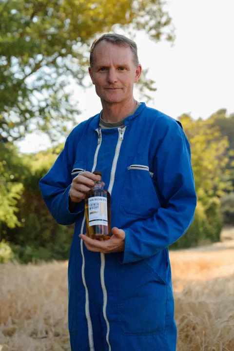 Portrait d'un agriculteur en bleu de travail, une bouteille de Soubeyran à la main, dans un champ d'orge